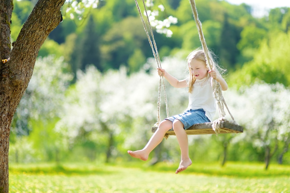 Cute little girl having fun on a swing in blossoming old apple tree garden outdoors on sunny spring day
